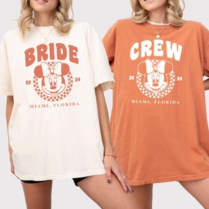 Puede incluir: Dos camisetas de gran tama&ntilde;o con un gr&aacute;fico inspirado en Mickey Mouse. La camiseta blanca dice "Bride" con el a&ntilde;o 20 y 24 a cada lado del gr&aacute;fico. La camiseta naranja dice "Crew" con el a&ntilde;o 20 y 24 a cada lado del gr&aacute;fico. Ambas camisetas dicen "Miami, Florida" debajo del gr&aacute;fico.