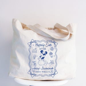 May include: Cream-colored tote bag with a blue graphic that reads "Happily Ever After" with a Minnie Mouse illustration. The bag also includes text that says "Ashley's Bachelorette" and "Disney World, FL". The bag has a simple design with a rectangular shape and two handles.