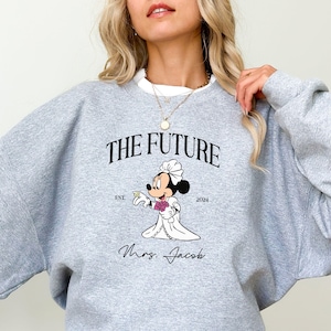 May include: Gray sweatshirt with a graphic of a cartoon bride Mickey Mouse holding a bouquet of flowers. The text "The Future" is above the graphic and "Est. 2004" is below. The text "Mrs. Jacob" is below the graphic.