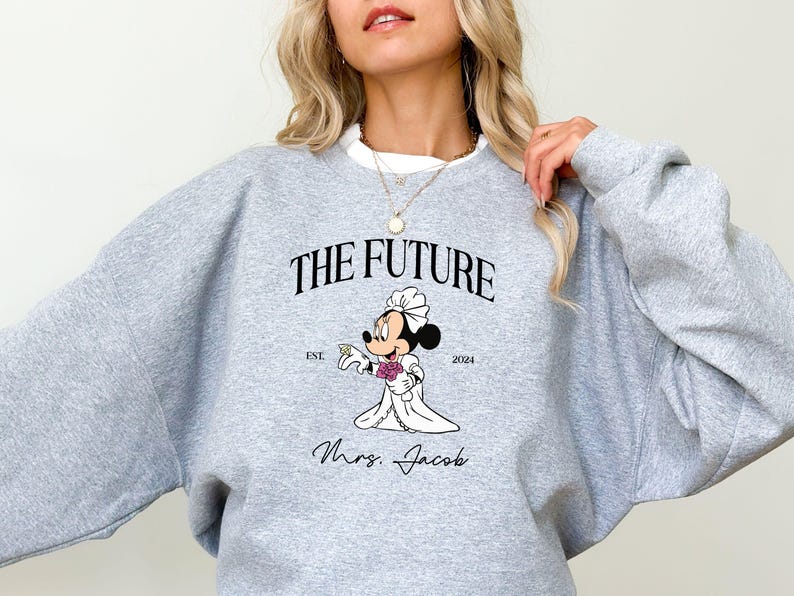 May include: Gray sweatshirt with a graphic of a cartoon bride Mickey Mouse holding a bouquet of flowers. The text "The Future" is above the graphic and "Est. 2004" is below. The text "Mrs. Jacob" is below the graphic.