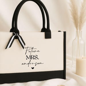 May include: A white canvas tote bag with black trim and handles. The bag has a black ribbon bow and the text "Future Mrs. Anderson" printed on the front.