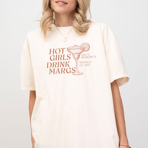 May include: A white t-shirt with the text "HOT GIRLS DRINK MARGS" in orange. The text is above a drawing of a margarita glass with a lime wedge. The text "EMILY'S BACHELORETTE NASHVILLE EST 2023" is printed in smaller orange text on the side of the glass.