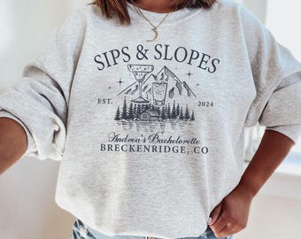 May include: Grey sweatshirt with the text "Sips & Slopes" and a graphic of two drinks, mountains, and trees. The text "Est. 2024" is below the graphic. The text "Andrea's Bachelorette Breckenridge, CO" is below the text.