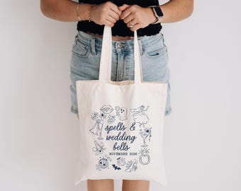 May include: A cream-coloured canvas tote bag featuring a dark blue graphic print of various icons including a ghost, a martini glass, a diamond ring, and a disco ball. The text reads spells & wedding bells and NOVEMBER 2026 in a dark blue font.