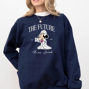 May include: A navy blue sweatshirt with a white graphic of Mickey Mouse dressed as a bride. The graphic includes the text "The Future" with the years "Est. 2024" and the text "Mrs. Jacob".