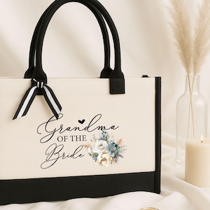 May include: Cream-colored tote bag with black trim and handles. The bag features the text "Grandma of the Bride" in elegant script, along with a floral design in blue and white. A black and white striped bow adorns the handle.