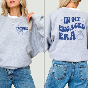 May include: Grey sweatshirt with the text "Future Mrs. Martinez" on the front and "In My Engaged Era" on the back. The sweatshirt has a ring and diamond graphic on the back.