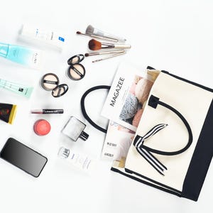 Op de afbeelding: Een witte canvas tote bag met zwarte bies en een zwart-wit gestreept lint. De tas is open en gevuld met verschillende items, waaronder make-upkwasten, een tijdschrift en een fles parfum.