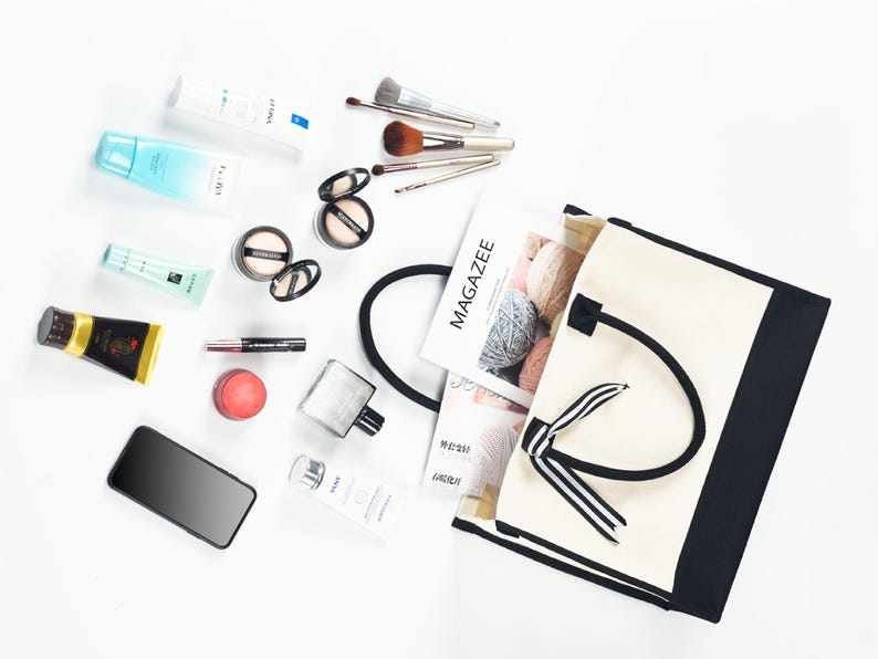 Op de afbeelding: Een witte canvas tote bag met zwarte bies en een zwart-wit gestreept lint. De tas is open en gevuld met verschillende items, waaronder make-upkwasten, een tijdschrift en een fles parfum.