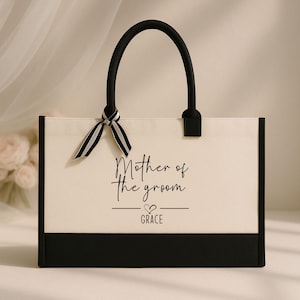 May include: A white canvas tote bag with black trim and handles. The bag has a black and white striped ribbon tied to the handles. The bag says "Mother of the groom - GRACE" in black text.