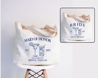 May include: Two white canvas tote bags with blue text and illustrations of cocktails. The first bag says "Bride Est. 2024 Wife of the Party". The second bag says "Maid of Honour Est. 2024 #justonemorecox Bride Squad".