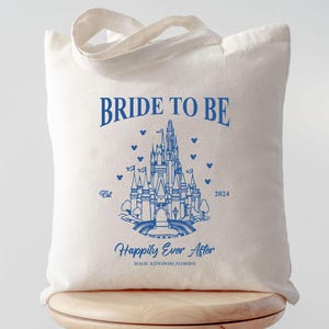 May include: A white tote bag with blue text that reads "Bride to Be" and a blue outline of Cinderella's castle. The text "Happily Ever After" is below the castle with the words "Magic Kingdom, Florida" underneath.