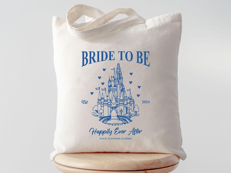 May include: A white tote bag with blue text that reads "Bride to Be" and a blue outline of Cinderella's castle. The text "Happily Ever After" is below the castle with the words "Magic Kingdom, Florida" underneath.