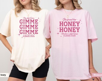 May include: Two oversized t-shirts, one ivory and one pink, with matching text in a dark pink color. The ivory shirt says "GIMME GIMME GIMME" and the pink shirt says "HONEY HONEY". Both shirts have the text "FLORIDA-2025".