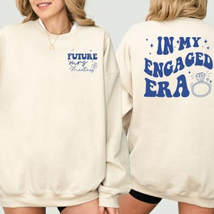May include: A beige sweatshirt with blue text that says "Future Mrs. Martinez" on the front and "In My Engaged Era" on the back. The text is in a retro font and includes a diamond ring graphic.