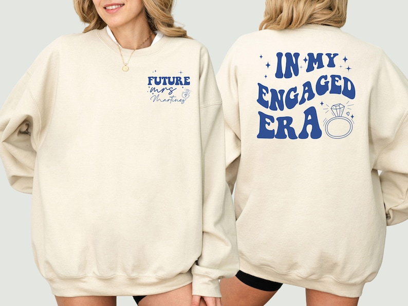 May include: A beige sweatshirt with blue text that says "Future Mrs. Martinez" on the front and "In My Engaged Era" on the back. The text is in a retro font and includes a diamond ring graphic.