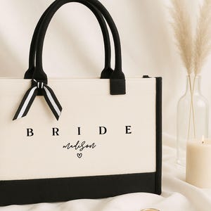 May include: A white canvas tote bag with black trim and handles. The bag has the word "BRIDE" printed in black letters and the name "madison" written in cursive with a heart symbol.