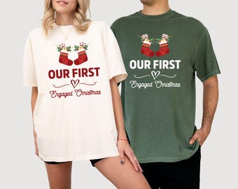 May include: Two t-shirts, one cream and one green, with the text "OUR FIRST" above a heart and the words "Engaged Christmas." Each shirt features a Christmas stocking design with candy canes and holly.