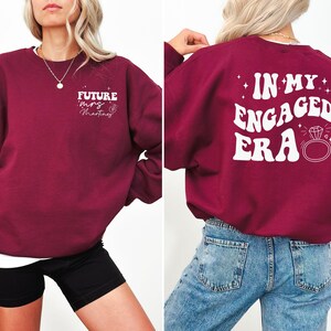 May include: Burgundy sweatshirt with white text that says "Future Mrs. Martinez" on the front and "In My Engaged Era" with a diamond ring on the back.