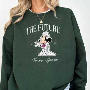 May include: A dark green sweatshirt with a white graphic of a cartoon mouse dressed as a bride. The text "The Future" is above the mouse and "Est. 2024" is below the mouse. The text "Mrs. Jacob" is below the mouse.