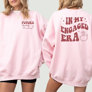 May include: Two pink sweatshirts with a red graphic print. One sweatshirt says "Future Mrs. Martinez" and the other says "In My Engaged Era" with a diamond ring graphic.