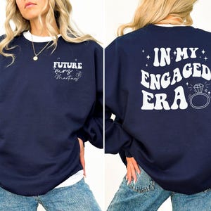 May include: Navy blue sweatshirt with white text on the front that says "Future Mrs. Martinez" and white text on the back that says "In My Engaged Era" with a diamond ring graphic.