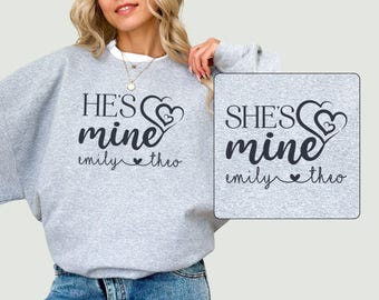 Op de afbeelding: Twee grijze sweatshirts met een hartvormig ontwerp en de tekst "He's mine" en "She's mine" met de namen "emily" en "theo" eronder.