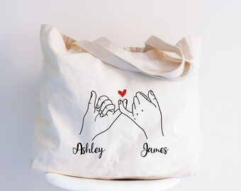 May include: White canvas tote bag featuring a black line drawing of two hands intertwined with a small red heart above them. The text "Ashley" and "James" is printed below the hands.