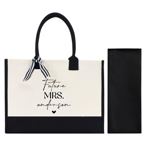 Op de afbeelding: Een witte canvas tote bag met zwarte afwerking en handvatten. De tas heeft een zwart lint om de handvatten gebonden en de tekst "Future Mrs. anderson" gedrukt op de voorkant. De tas wordt geleverd met een bijpassende zwarte ritszak.