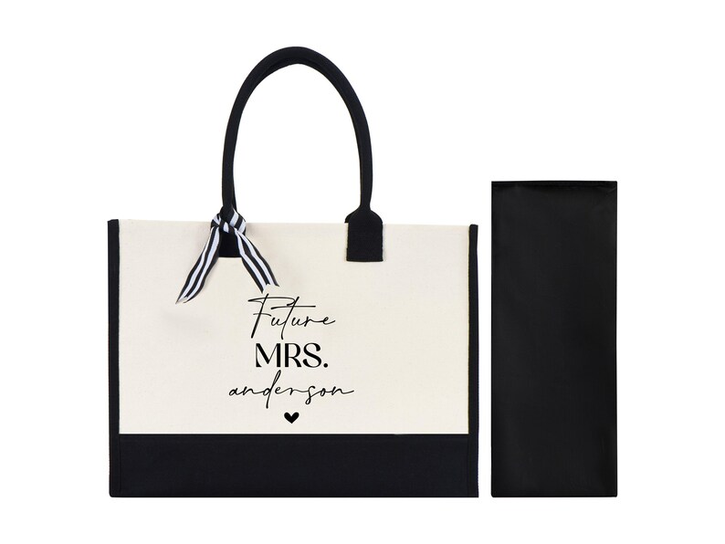 Op de afbeelding: Een witte canvas tote bag met zwarte afwerking en handvatten. De tas heeft een zwart lint om de handvatten gebonden en de tekst "Future Mrs. anderson" gedrukt op de voorkant. De tas wordt geleverd met een bijpassende zwarte ritszak.