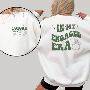 May include: White sweatshirt with green text that says "In My Engaged Era" with a diamond ring graphic. The back of the sweatshirt has green text that says "Future Mrs. Martinez" with a ring graphic.