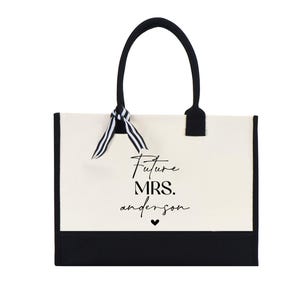 Op de afbeelding: Een witte canvas tote bag met zwarte bies en handvatten. De tas heeft een zwart-wit gestreept lint dat om de handvatten is gebonden. De tas heeft de tekst "Future Mrs. anderson" in zwart op de voorkant gedrukt.