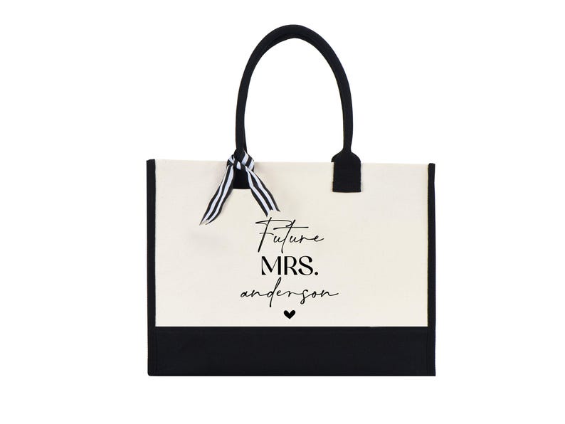 Op de afbeelding: Een witte canvas tote bag met zwarte bies en handvatten. De tas heeft een zwart-wit gestreept lint dat om de handvatten is gebonden. De tas heeft de tekst "Future Mrs. anderson" in zwart op de voorkant gedrukt.