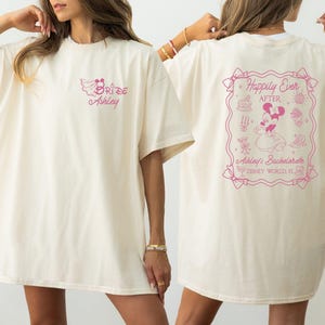 May include: Cream-colored t-shirt with pink text and graphics. The front features "Bride Ashley" in script. The back displays a framed design with "Happily Ever After," Minnie Mouse, and "Ashley's Bachelorette Disney World FL."