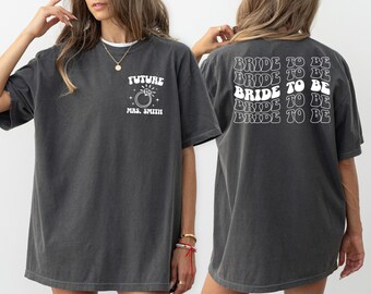 May include: Two charcoal grey t-shirts with white text. The front shirt says "FUTURE MRS. SMITH" with a stylised ring and diamond. The back shirt says "BRIDE TO BE" five times in a retro font.