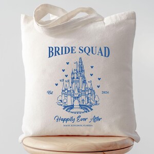 May include: A white canvas tote bag with blue text that reads "Bride Squad" and a blue outline of Cinderella's castle. The text "Happily Ever After" is below the castle with the words "Magic Kingdom, Florida" underneath.
