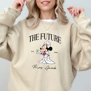 May include: Beige sweatshirt with a graphic of a cartoon bride Mickey Mouse holding a bouquet of flowers. The text "The Future" is above the graphic and "Est. 2024" is below. The text "Mrs. Jacob" is below the graphic.