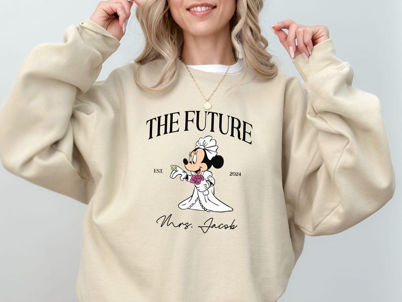 May include: Beige sweatshirt with a graphic of a cartoon bride Mickey Mouse holding a bouquet of flowers. The text "The Future" is above the graphic and "Est. 2024" is below. The text "Mrs. Jacob" is below the graphic.