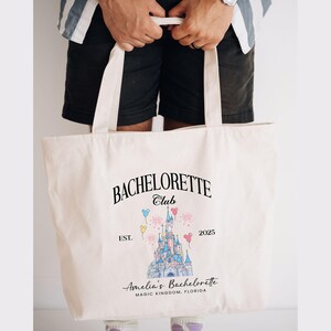 May include: A white canvas tote bag with a black graphic design featuring a cartoon castle, balloons, and the text "Bachelorette Club Est. 2025 Amelia's Bachelorette Magic Kingdom, Florida".