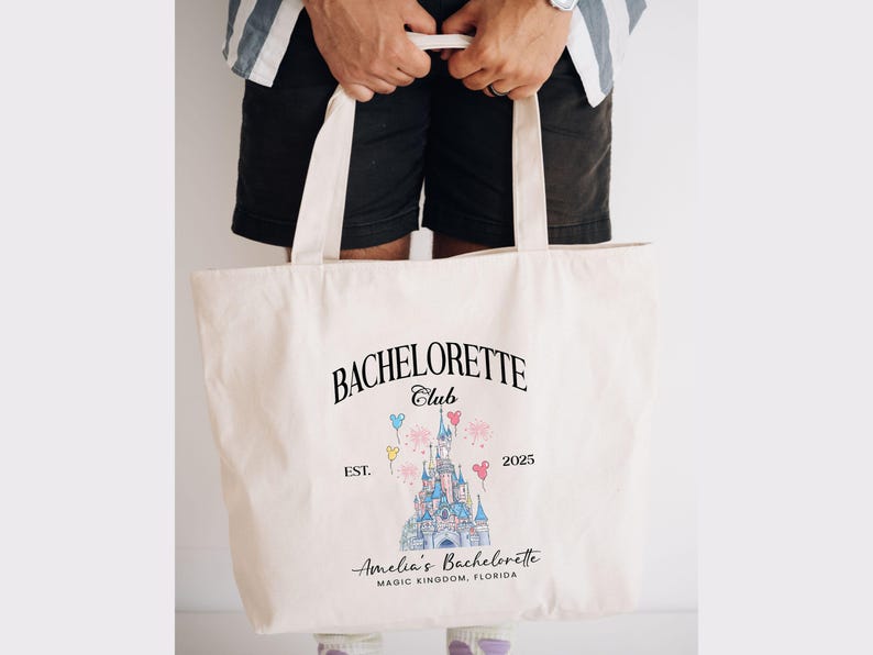 May include: A white canvas tote bag with a black graphic design featuring a cartoon castle, balloons, and the text "Bachelorette Club Est. 2025 Amelia's Bachelorette Magic Kingdom, Florida".