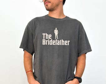 May include: A dark grey t-shirt with a white graphic of a man in a suit and the text "The Bridefather".
