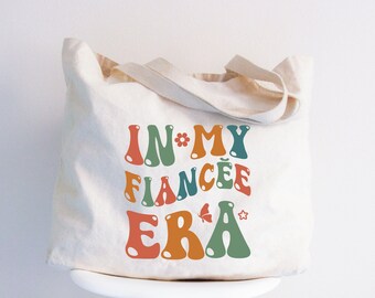 May include: Cream-colored tote bag with the text "IN MY FIANCÉE ERA" in retro-style, colorful lettering. The design includes a flower, butterfly, and star. The bag has a simple, neutral design with matching handles.