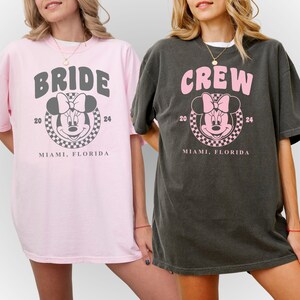 Puede incluir: Dos camisetas de color rosa y gris con un gr&aacute;fico inspirado en Mickey Mouse. Una camiseta dice "Bride" y la otra dice "Crew". Ambas camisetas dicen "Miami, Florida" y "20 24" debajo del gr&aacute;fico.