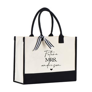 Op de afbeelding: Een witte canvas tote bag met zwarte bies en handvatten. De tas heeft een zwart-wit lint om de handvatten gebonden. De tas heeft de tekst "Future Mrs. Anderson" in het zwart op de voorkant gedrukt.
