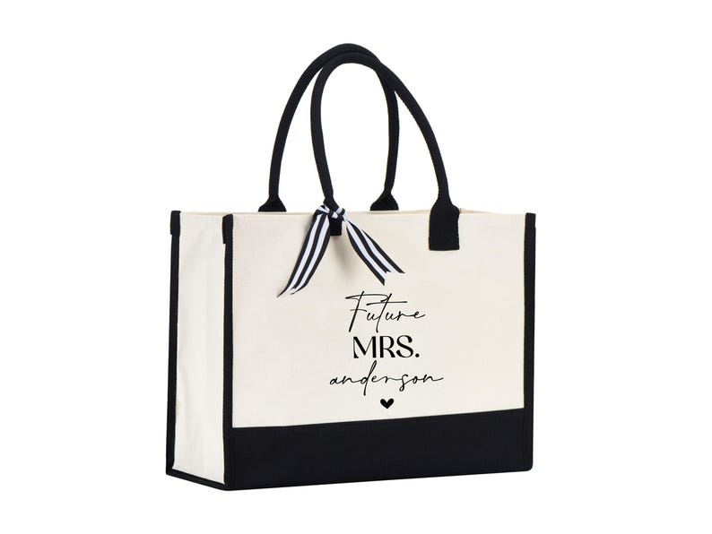 Op de afbeelding: Een witte canvas tote bag met zwarte bies en handvatten. De tas heeft een zwart-wit lint om de handvatten gebonden. De tas heeft de tekst "Future Mrs. Anderson" in het zwart op de voorkant gedrukt.