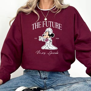May include: Burgundy sweatshirt with a white graphic of Mickey Mouse dressed as a bride. The graphic says "The Future" with "Est. 2024" and "Mrs. Jacob" below the image.