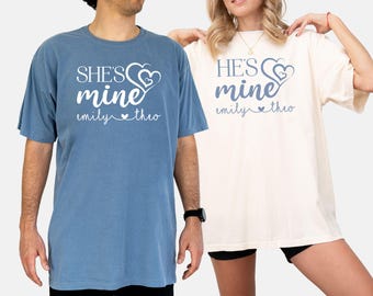 Op de afbeelding: Twee T-shirts met een ontwerp voor een stel. Het blauwe shirt zegt "SHE'S mine emily ♥ theo" en het witte shirt zegt "HE'S mine emily ♥ theo".