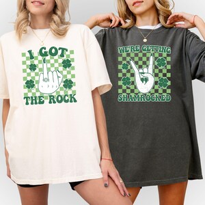 May include: Two oversized t-shirts, one cream and one charcoal, each with a graphic design. The cream shirt says "I GOT THE ROCK" with a hand gesture and shamrocks. The charcoal shirt says "WE'RE GETTING SHAMROCKED" with a hand gesture and shamrocks.
