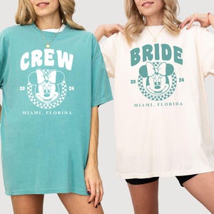 Puede incluir: Dos camisetas de gran tama&ntilde;o con un gr&aacute;fico de Mickey Mouse y texto. La camiseta azul claro dice "CREW" y la camiseta blanca dice "BRIDE". Ambas camisetas tienen el texto "Miami, Florida" y los n&uacute;meros "20" y "24" impresos en ellas.