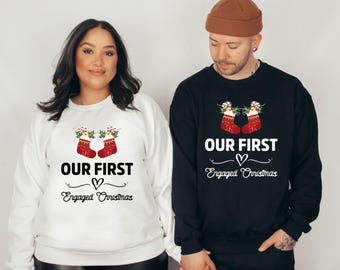 May include: Matching white and black sweatshirts with the text "OUR FIRST Engaged Christmas". Each sweatshirt features a design of two red Christmas stockings with holly and candy canes. A heart symbol is between the words.